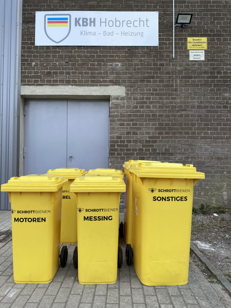 Das Bild zeigt einige Tonnen zur Metalltrennung vor dem Firmenlogo des Partnerunternehmens.
