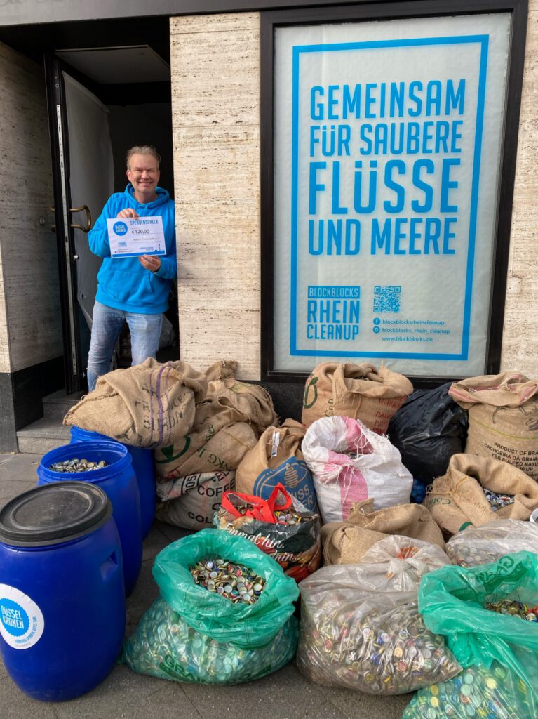 Das Bild zeigt einen Helfer mit dem Spendenscheck in der Hand neben den gesammelten Kronkorken.