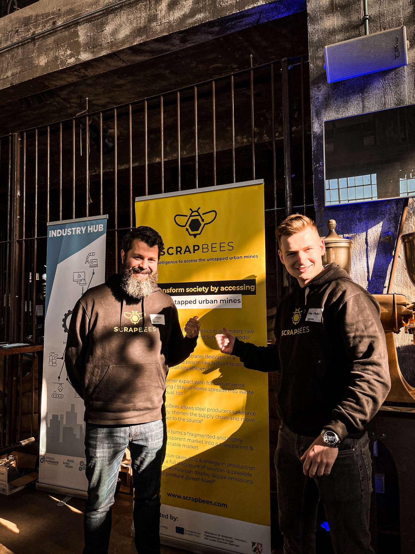 Das Bild zeigt zwei Mitarbeiter der ScrapBees GmbH mit einem Rollup zum Startup.