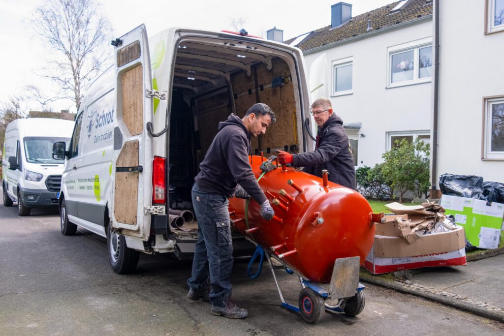 Das Bild zeigt zwei Fahrer der SchrottBienen dabei, wie sie einen schweren Kessel mit Hilfe von Sackkarre und Spanngurt in ihr Fahrzeug einladen.