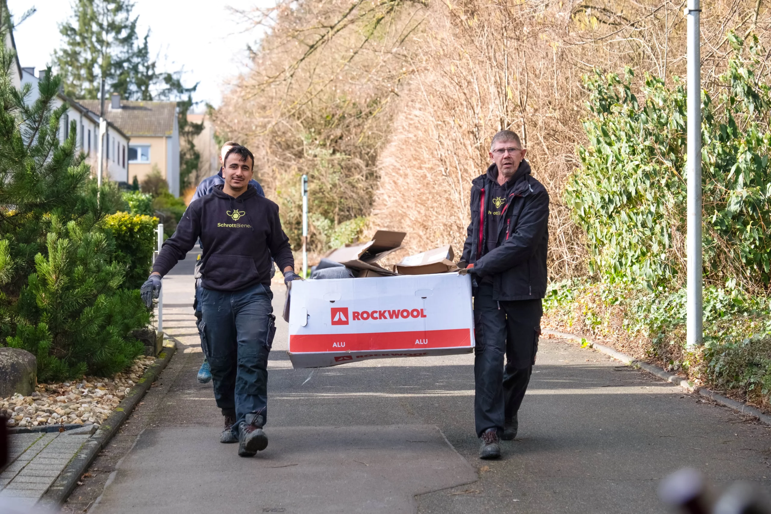 Das Bild zeigt zwei Fahrer der SchrottBienen dabei, wie sie gemeinsam einen Karton in Richtung Fahrzeug tragen.