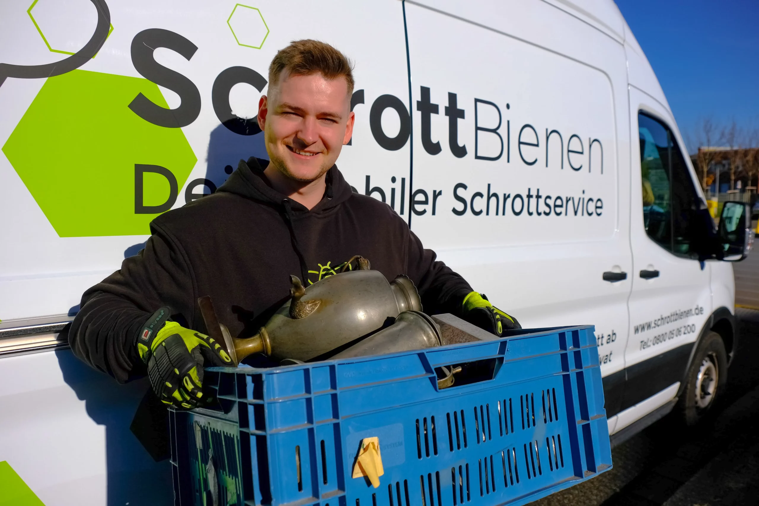 Das Bild zeigt einen Fahrer der SchrottBienen dabei, wie er eine Kiste voller Zinn in den Händen hält. Im Hintergrund ist ein Firmenfahrzeug der SchrottBienen zu sehen.