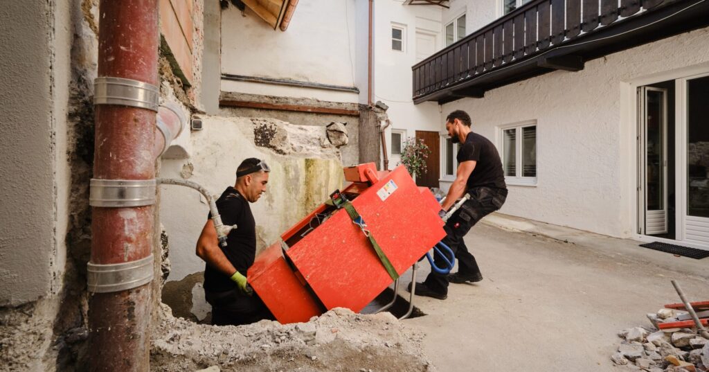 Zwei Mitarbeiter der SchrottBienen tragen eine alte rote Heizungsanlage aus einem Keller – Tragehilfe beim Heizungstausch.